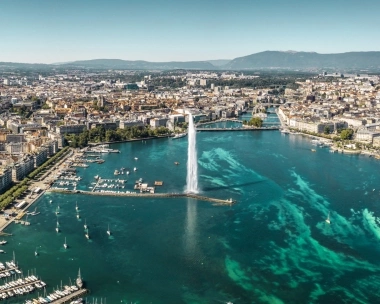 View of Geneva with the Jet d'Eau fountain in the foreground and Lake Geneva, surrounded by buildings and mountains in the background.