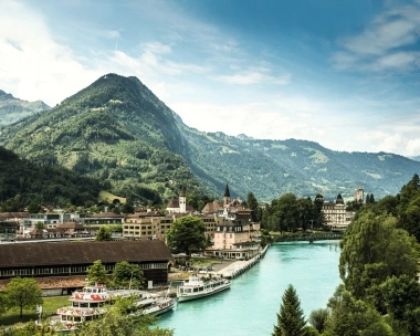 Stadtansicht mit Fluss, Booten und umliegenden Bergen im Hintergrund.