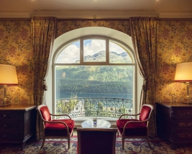 Stanza accogliente con due poltrone rosse, un tavolo e una grande finestra con vista su montagne e lago.