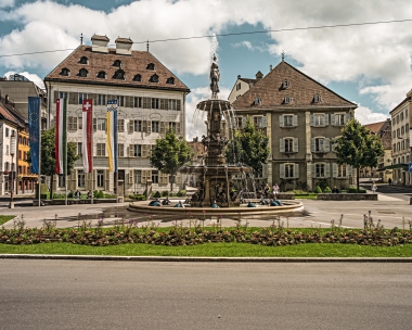 Eine hübsche Altstadt mit einem historischen Brunnen im Zentrum und europäischen Landesflaggen davor.