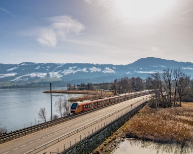 A regional train travels along a road by the lake shore, with snow-covered mountains in the background.