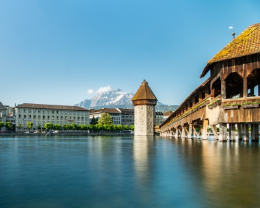 An old wooden bridge and a tower over a calm river, with snow-capped mountains in the background.