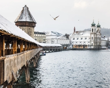 Wooden bridge over a river with a tower and snow-covered buildings in the background.