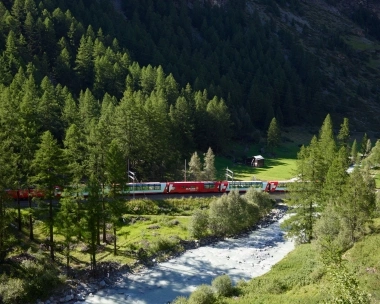 Train rouge traversant une vallée boisée avec une rivière au premier plan.