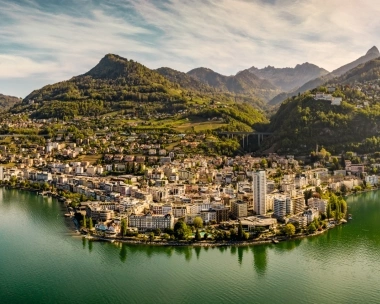 Città sul lago con colline boscose e montagne sullo sfondo.