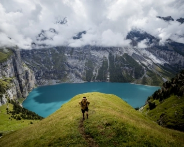 A person hikes on a green hill with a turquoise mountain lake in the background.