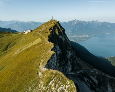 Cresta montuosa con torre di trasmissione, pendio ripido e ampia vista su una valle con lago e montagne sullo sfondo.