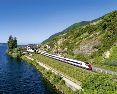 Train traveling along a lake with houses and green hills in the background.