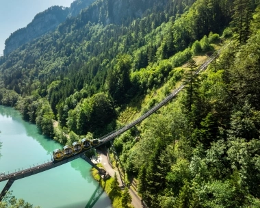 Una funicolare su un viadotto sopra un fiume turchese in un paesaggio montano boscoso.