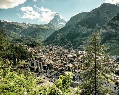 View of an alpine valley with a town, surrounded by green mountains and a prominent mountain peak in the background.