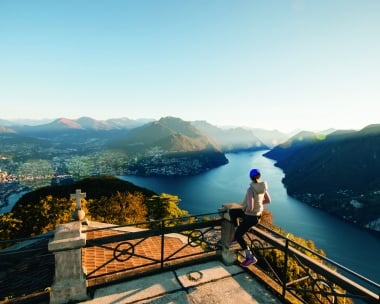 Frau überblickt von einer Terrasse auf einem Berg eine See und Berglandschaft.
