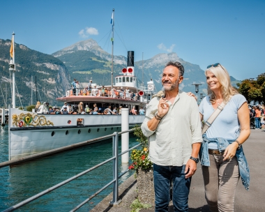 A steamship station on a lake with mountains in the background and two people walking along the shore.