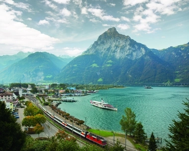 Mountain landscape with lake, train, and ship in the foreground.