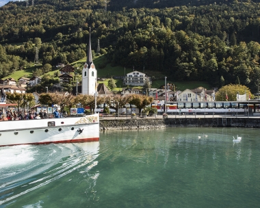 A ferry on a lake, with a church and houses in a green mountain landscape in the background.