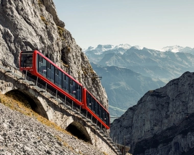 Un train à crémaillère rouge circule sur une section escarpée devant une paroi rocheuse, avec des montagnes en arrière-plan.
