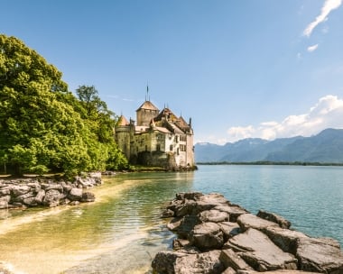 Ein mittelalterliches Schloss direkt am See, im Hintergrund die Berge, bei sonnigem Wetter.