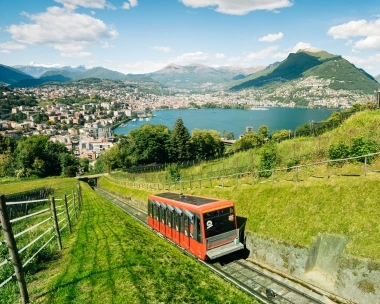Eine rote Standseilbahn fährt durch grüne Wiesen mit einer Stadt, einem See und Bergen im Hintergrund.