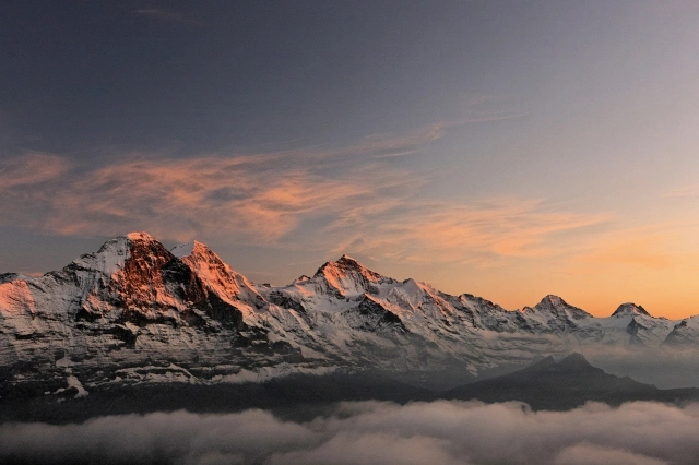 Eine Bergkette bei Sonnenaufgang mit schneebedeckten Gipfeln und leicht bewölktem Himmel.